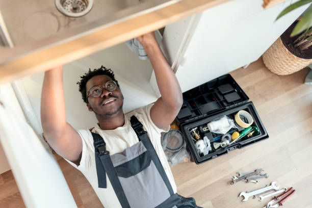 Person in overalls assembles furniture underneath a wooden table, with tools nearby.