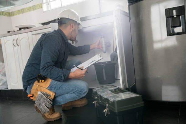 Technician crouches beside a refrigerator, inspecting it with a clipboard in a kitchen.