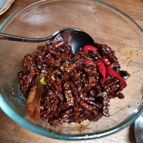 Een schaaltje tempeh in een glazen kom, donkerbruin met rode chilipepers en een lepel op een houten ondergrond.