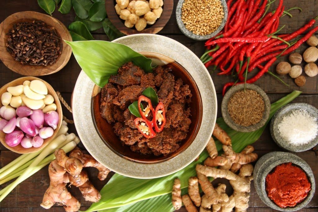 Een gerecht met rundvleesrendang, omringd door kruiden en ingredi&euml;nten, op een houten ondergrond.