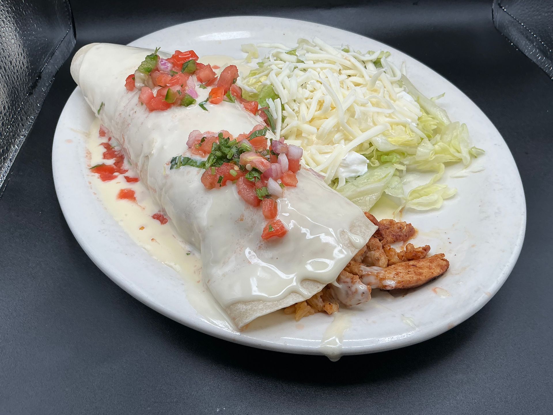 Burrito smothered in white sauce, topped with salsa, served with shredded cheese and lettuce on a white plate.