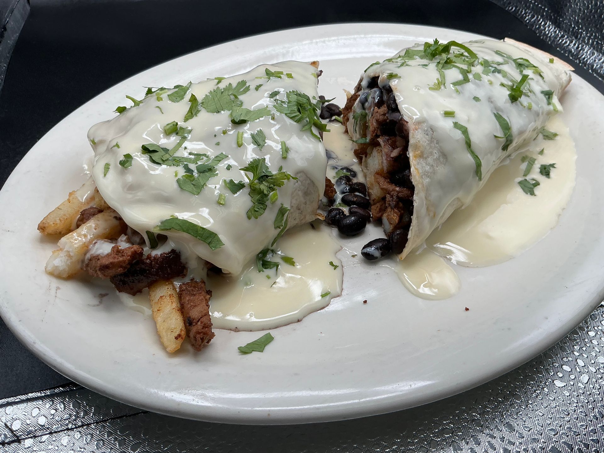 Burrito covered in creamy white sauce, with black beans, and chopped cilantro, served with fries.
