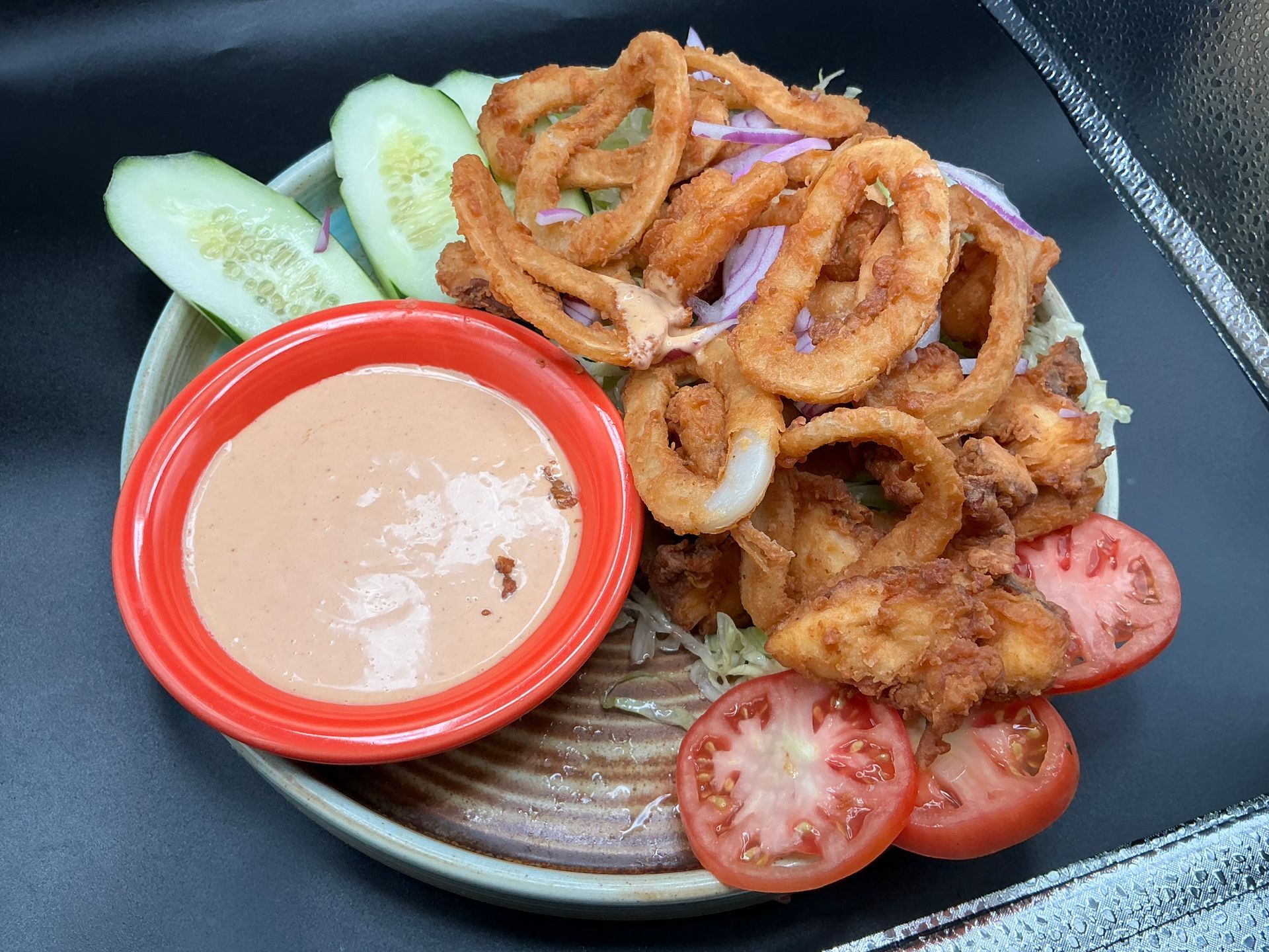 Fried calamari and shrimp with sauce, tomatoes, cucumber, and onions on a plate.