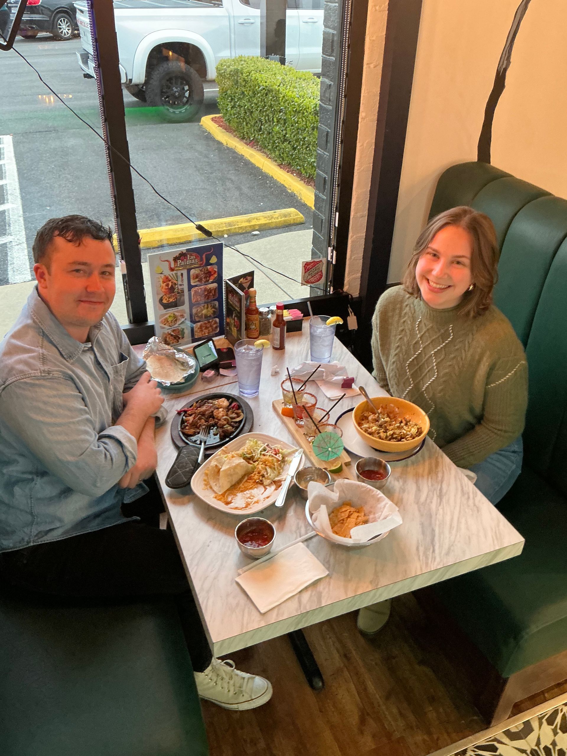 Two people seated at a restaurant table with food, smiling. Green booth, large window, and brick exterior.