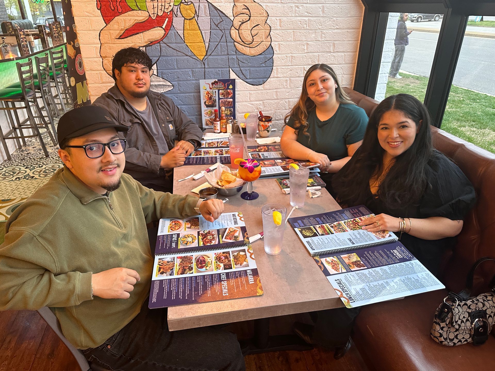 Four people seated at a table in a restaurant, looking at menus. Colorful mural in background.