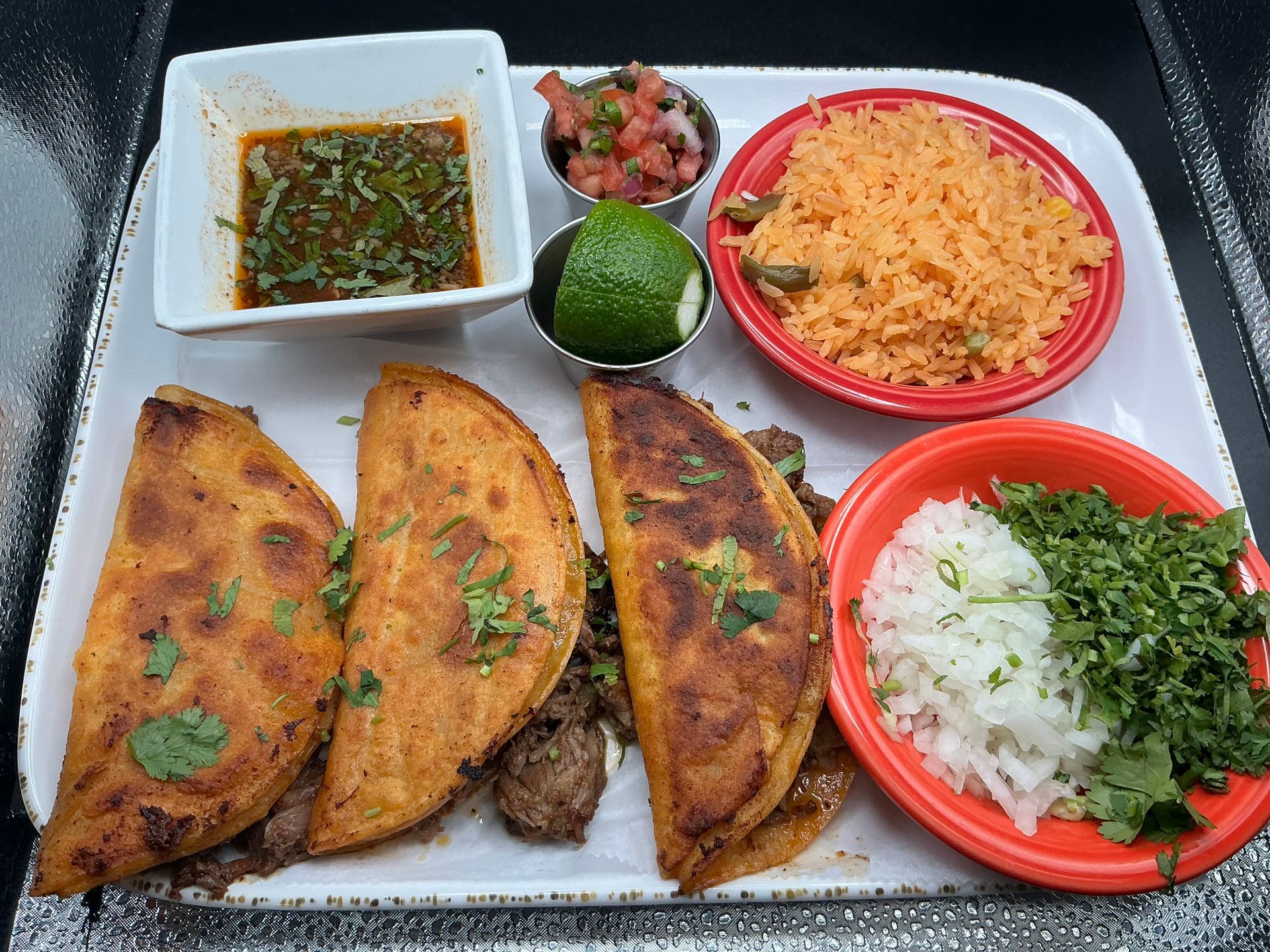 Birria tacos with salsa, rice, onions, cilantro, and lime on a platter.