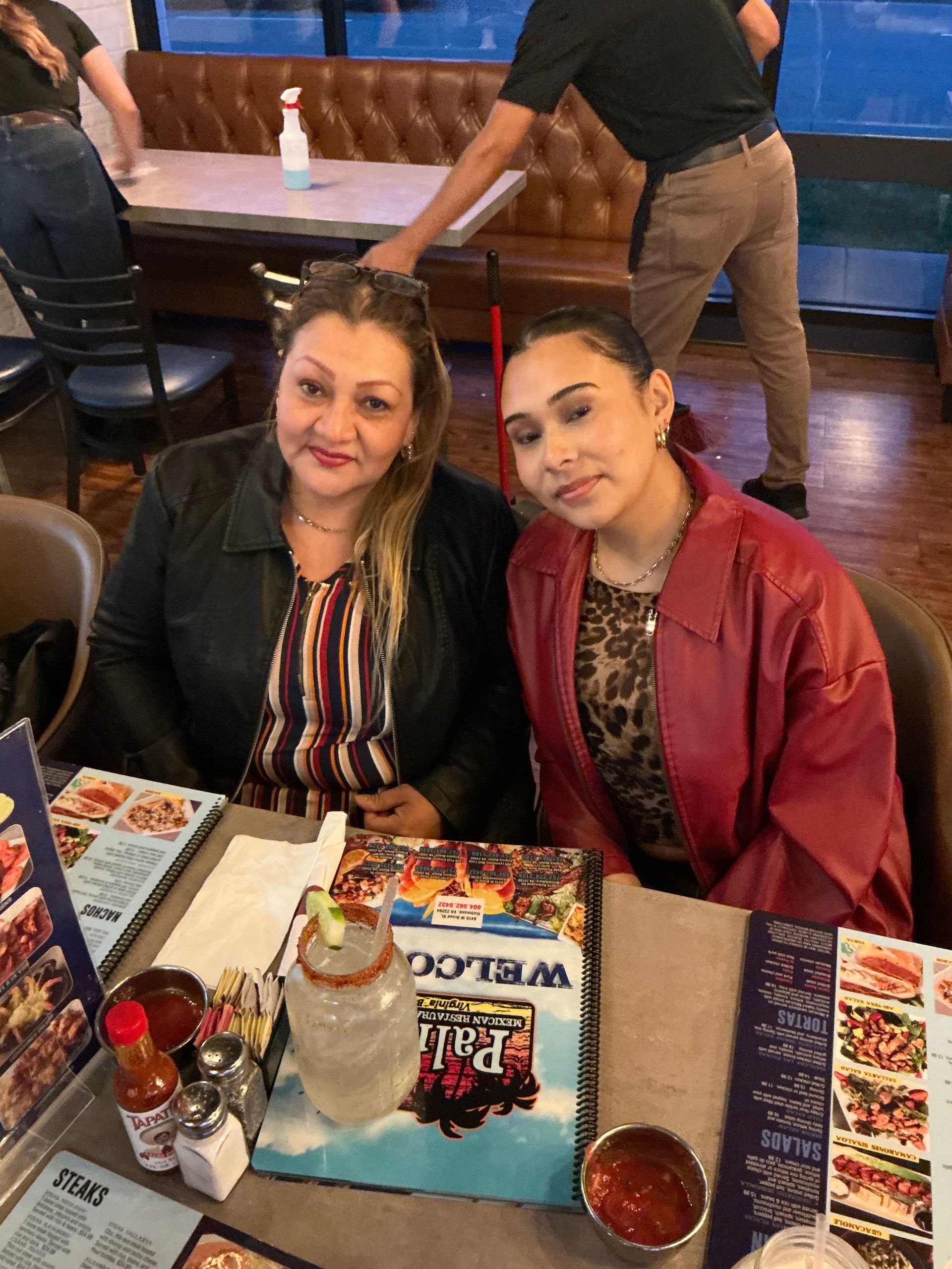 Two women at a restaurant table. One in a red jacket, the other with a striped shirt. Menus and food are visible.