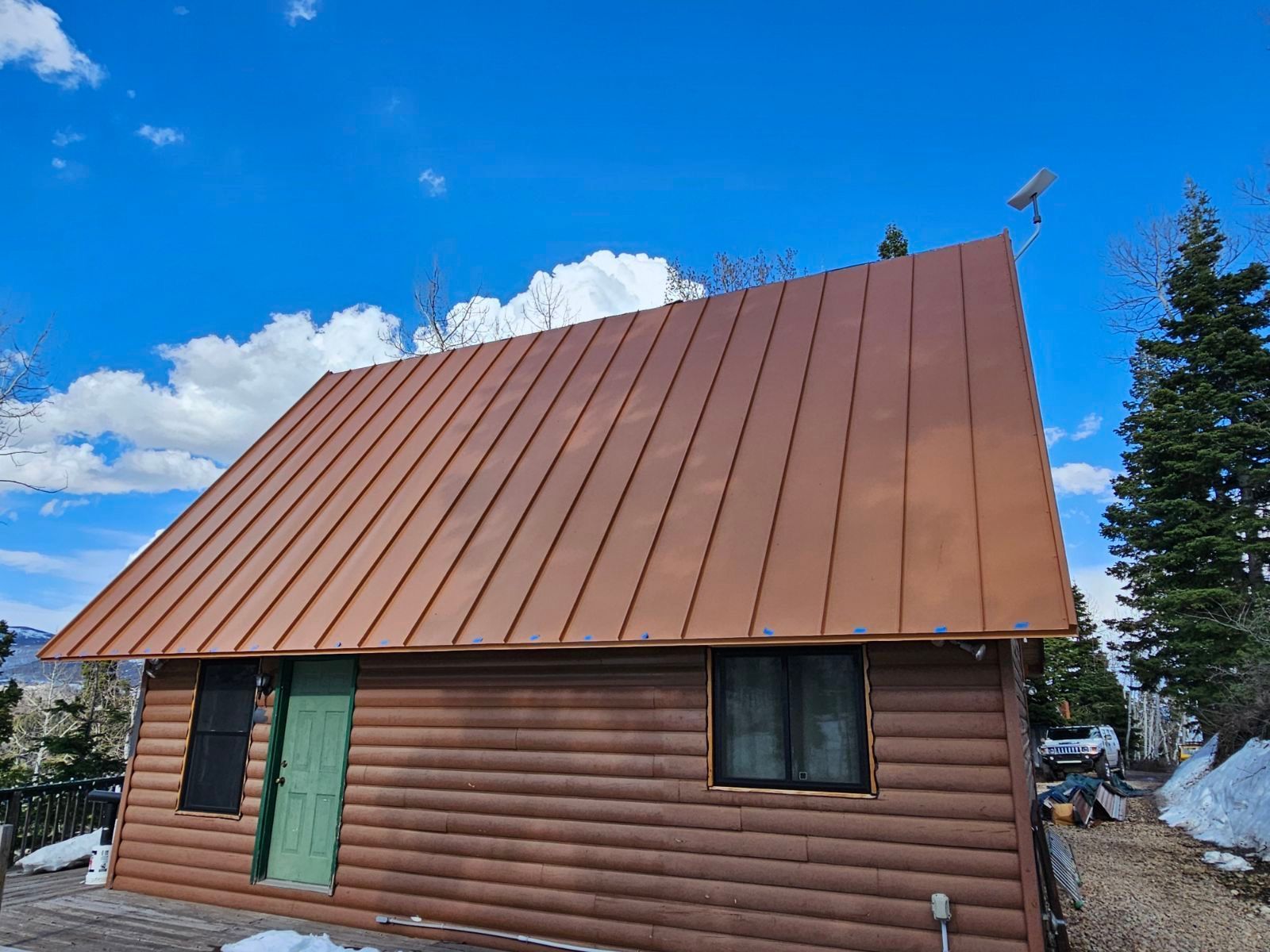 Log cabin with brown metal roof against a blue sky, with snow and trees in view.
