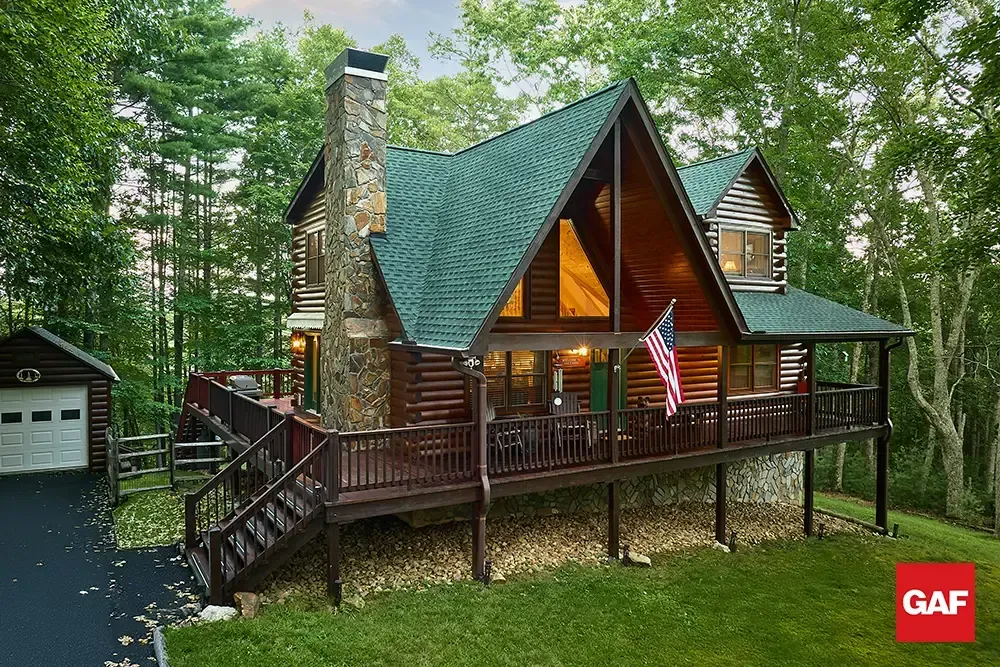 Rustic log cabin with green roof and wraparound deck nestled in a wooded setting