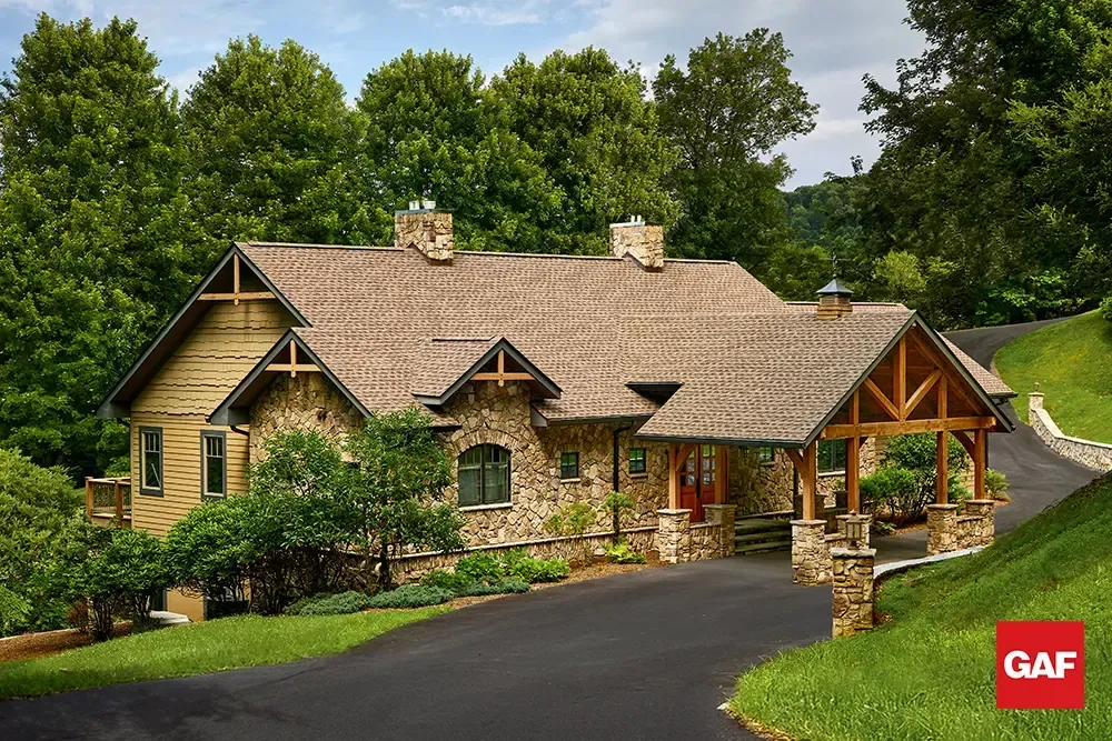 Stone-and-wood house with gabled roof, stone chimney, and driveway surrounded by green trees.
