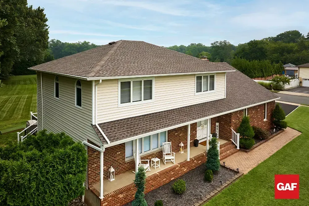 Two-story beige house with brown shingle roof, white trim, and front porch in a green suburban setting