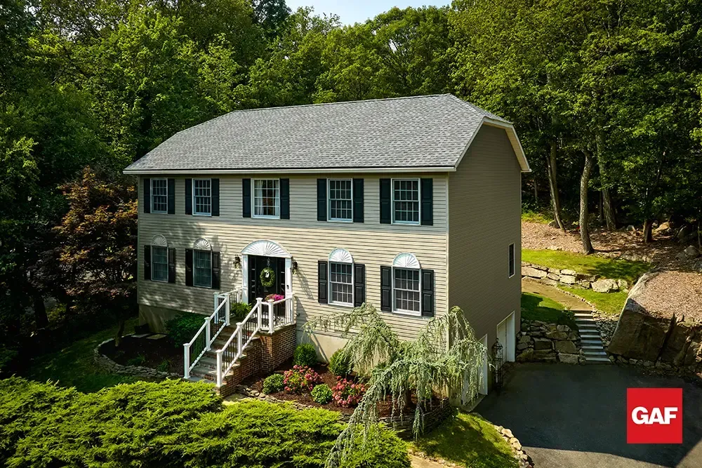 Two-story tan house with white trim and green shutters, surrounded by trees and a landscaped front yard