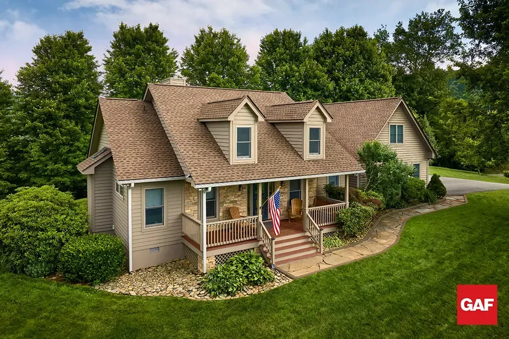 Suburban house with tan siding, shingled roof, front porch, and an American flag surrounded by green lawn and trees
