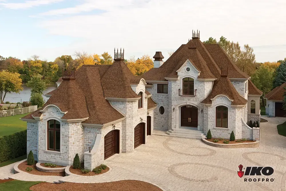 Stone Tudor-style house with brown shingle roofs, arched garage, and circular driveway at sunset