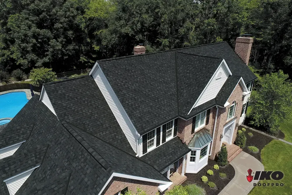 Aerial view of a house with dark roof, white trim, and a blue pool beside dense trees.