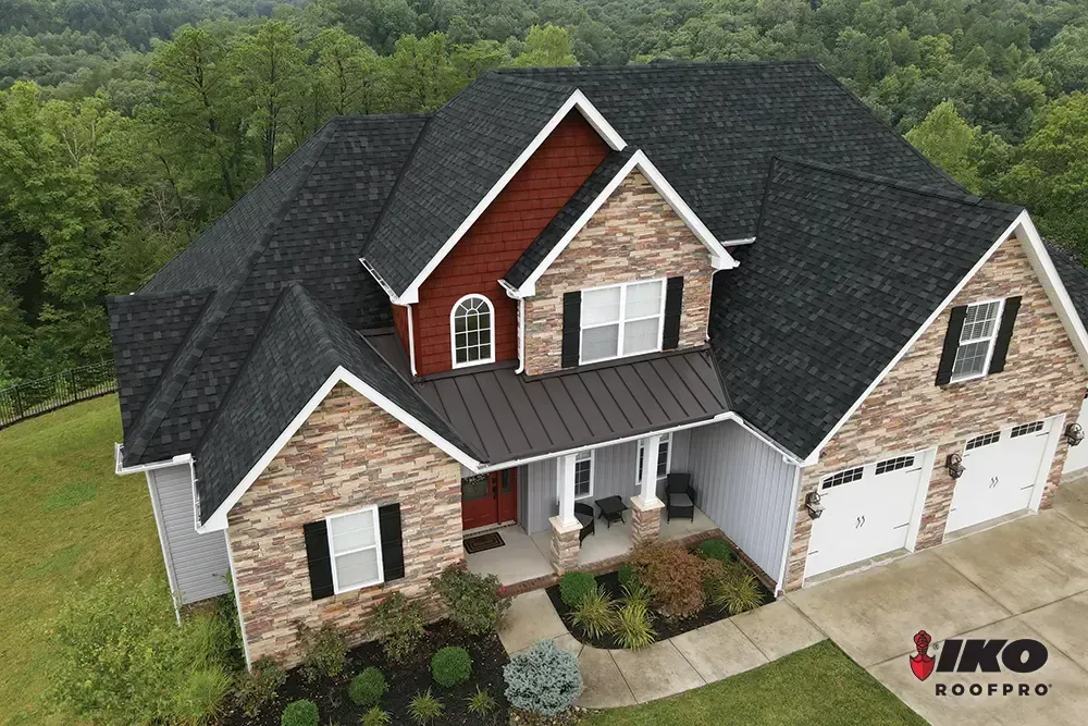 Aerial view of a brick-and-siding suburban house with a dark roof and front porch, surrounded by trees.