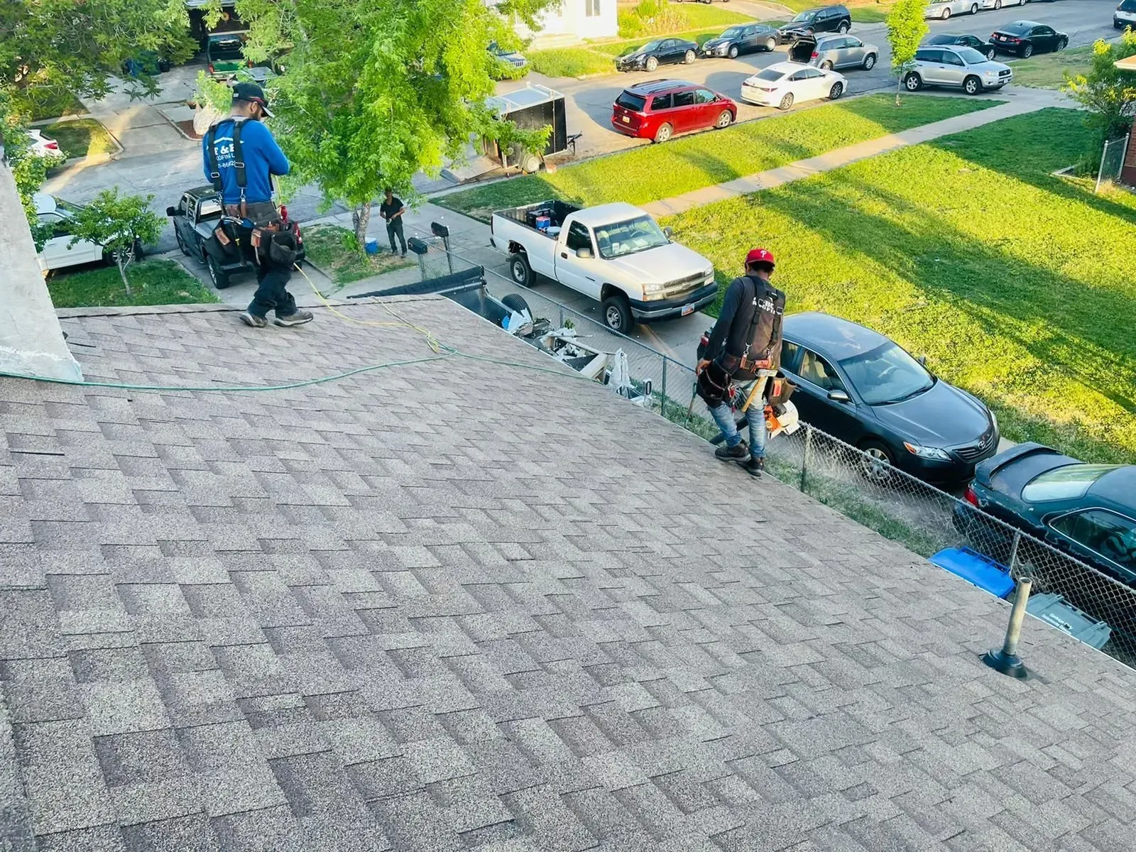 Roofers working on a shingled roof, with parked vehicles and a street below.