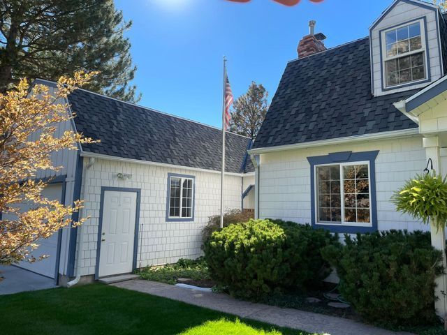 White house exterior with dark shingled roofs, dormer windows, shrubs, and an American flag under a blue sky