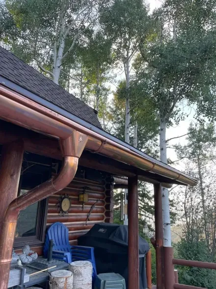 Copper gutters on a cabin roof with trees in the background. Deck with chairs, a grill, and firewood.