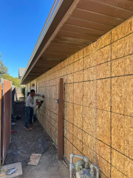 Two people installing wood siding on a building wall under a brown roof, outdoors.