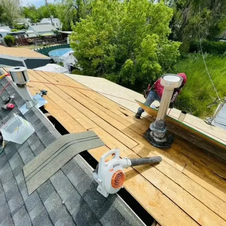 Roofer uses a blower and works on a partially shingled roof with a tall vent, surrounded by trees.