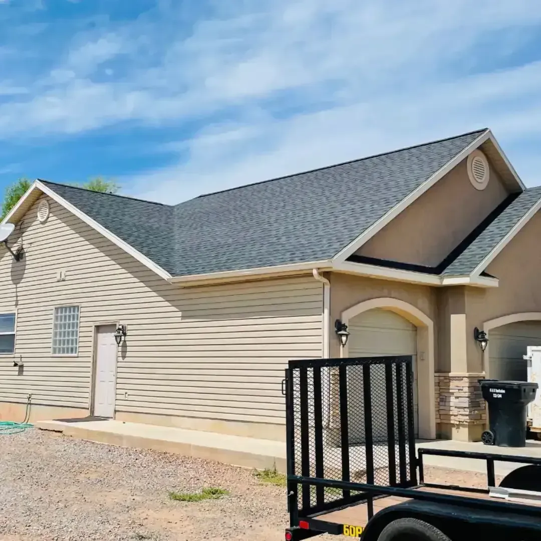 Tan house with gray roof and light blue sky. Black trailer in the foreground.