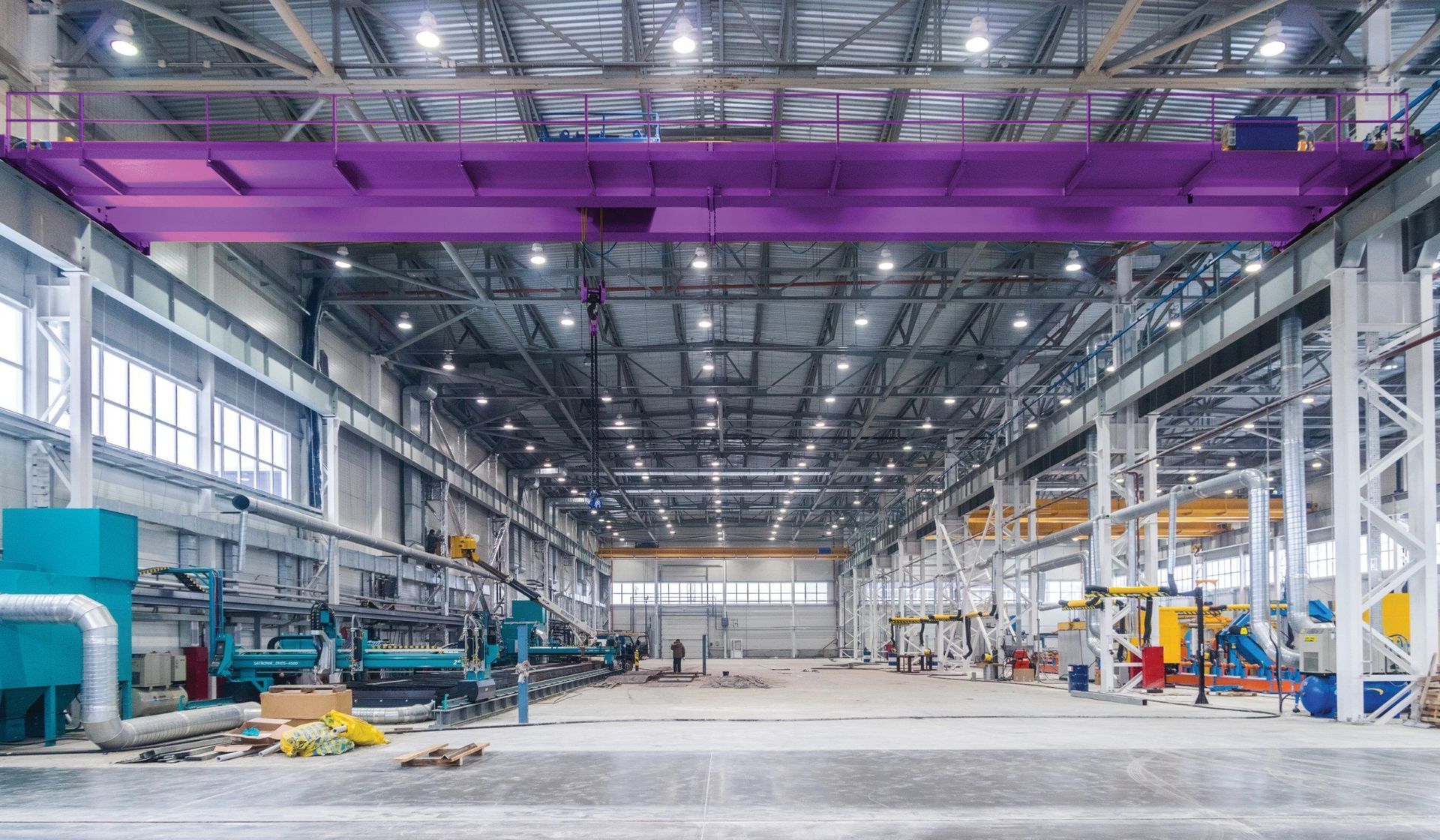 Purple overhead crane in a large industrial warehouse; machinery and equipment line the floor.