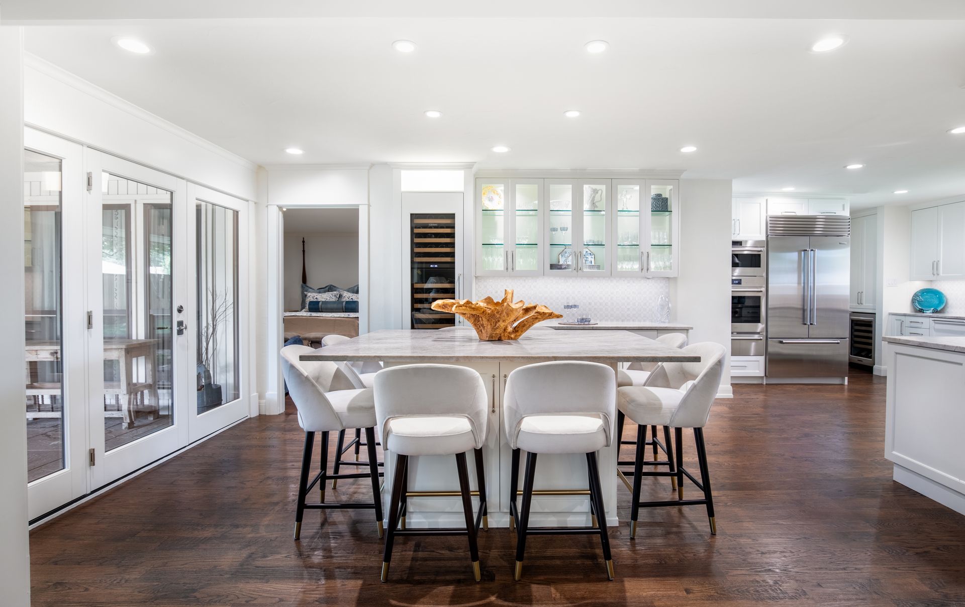 A modern, white kitchen with a central island featuring four bar stools, dark wood floors, and stainless steel appliances.