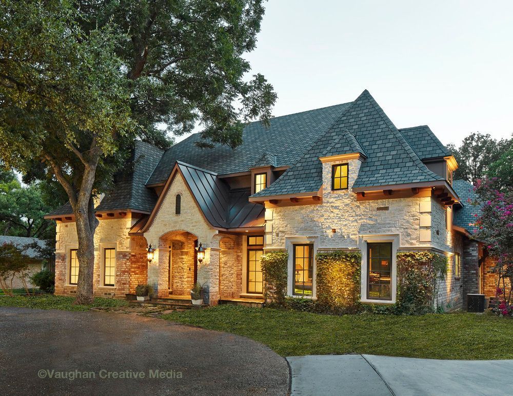 Stone and dark roofed house with a curving driveway, surrounded by greenery. Richardson exterior renovation.