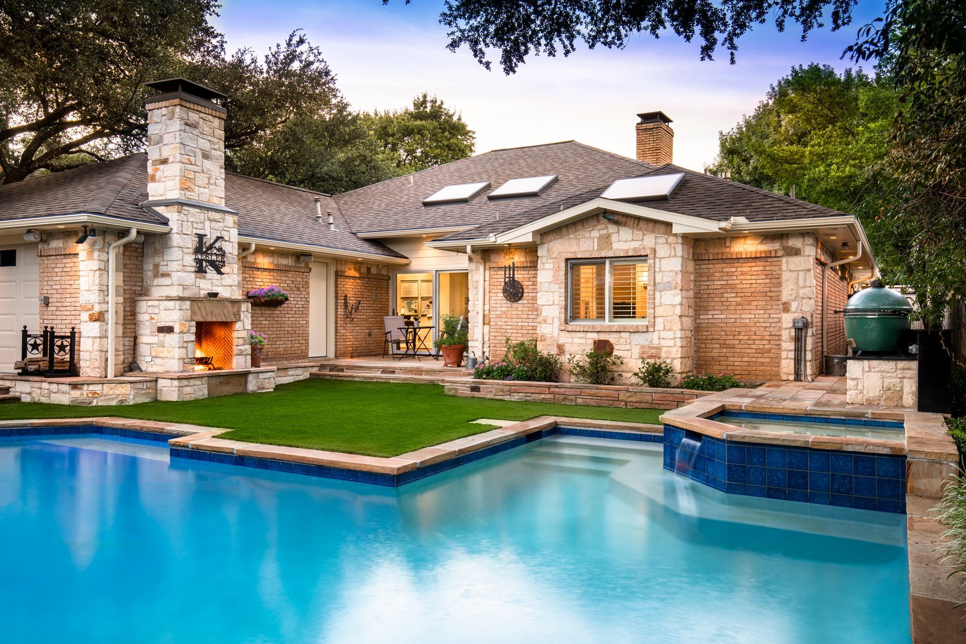 Backyard with a swimming pool, attached hot tub, manicured lawn, and a stone house with an outdoor fireplace.
