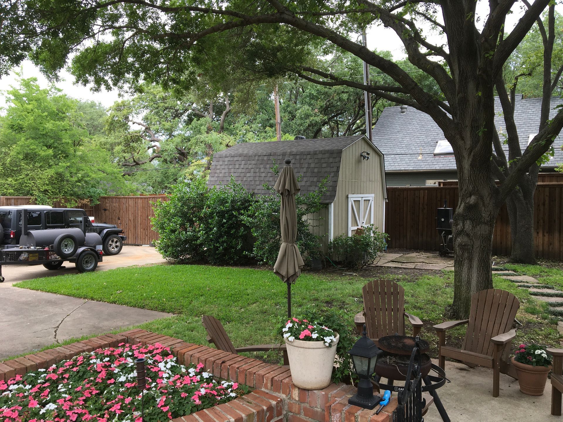 Backyard patio with potted flowers, wooden chairs, and a shaded tree beside a lawn and driveway