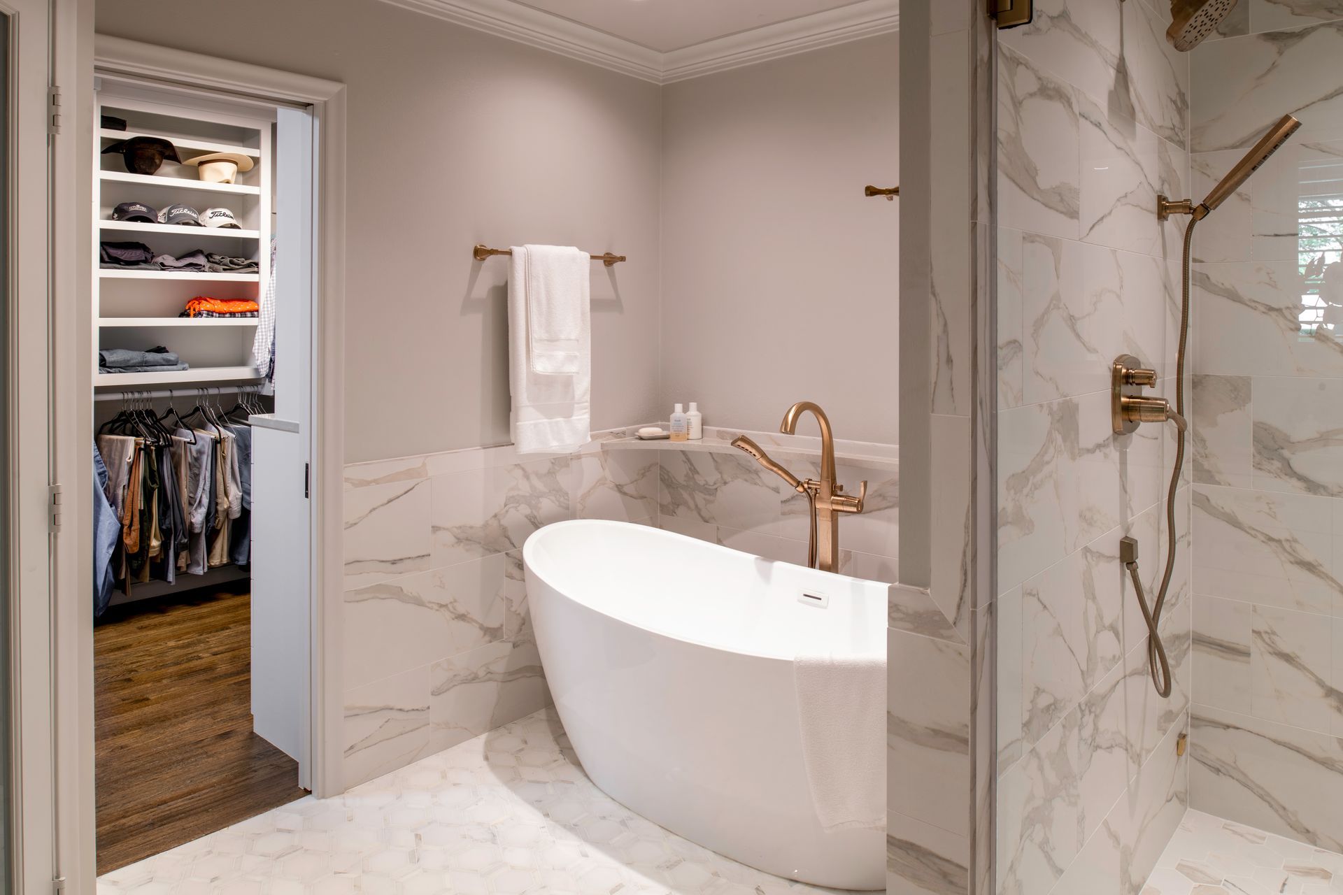 Luxury bathroom with marble walls, freestanding white tub, gold fixtures, and open closet in the background