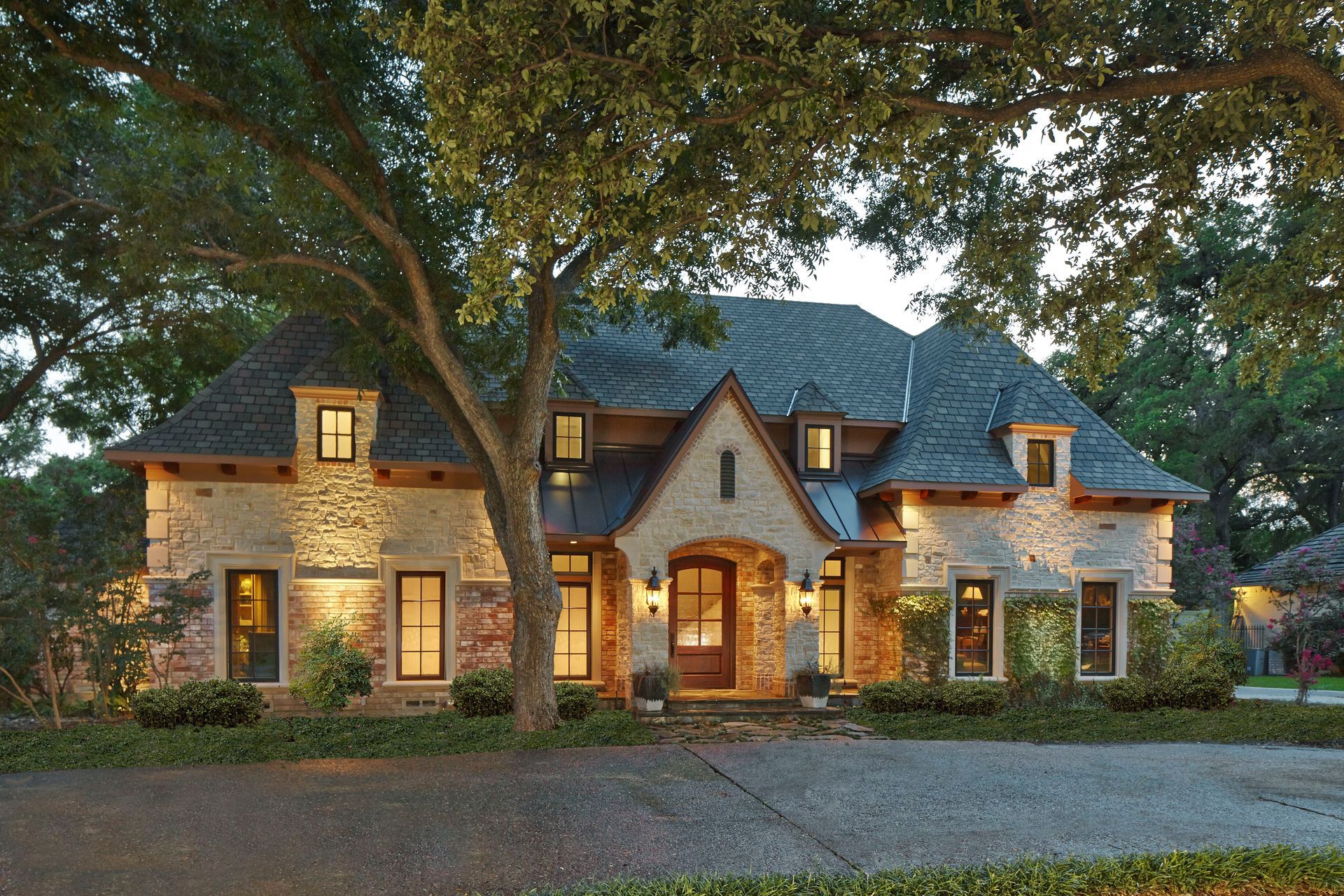 Stonefaced house with dormers, lit warmly at dusk, fronted by a driveway and large tree. Northwood Hills Exterior renovation.