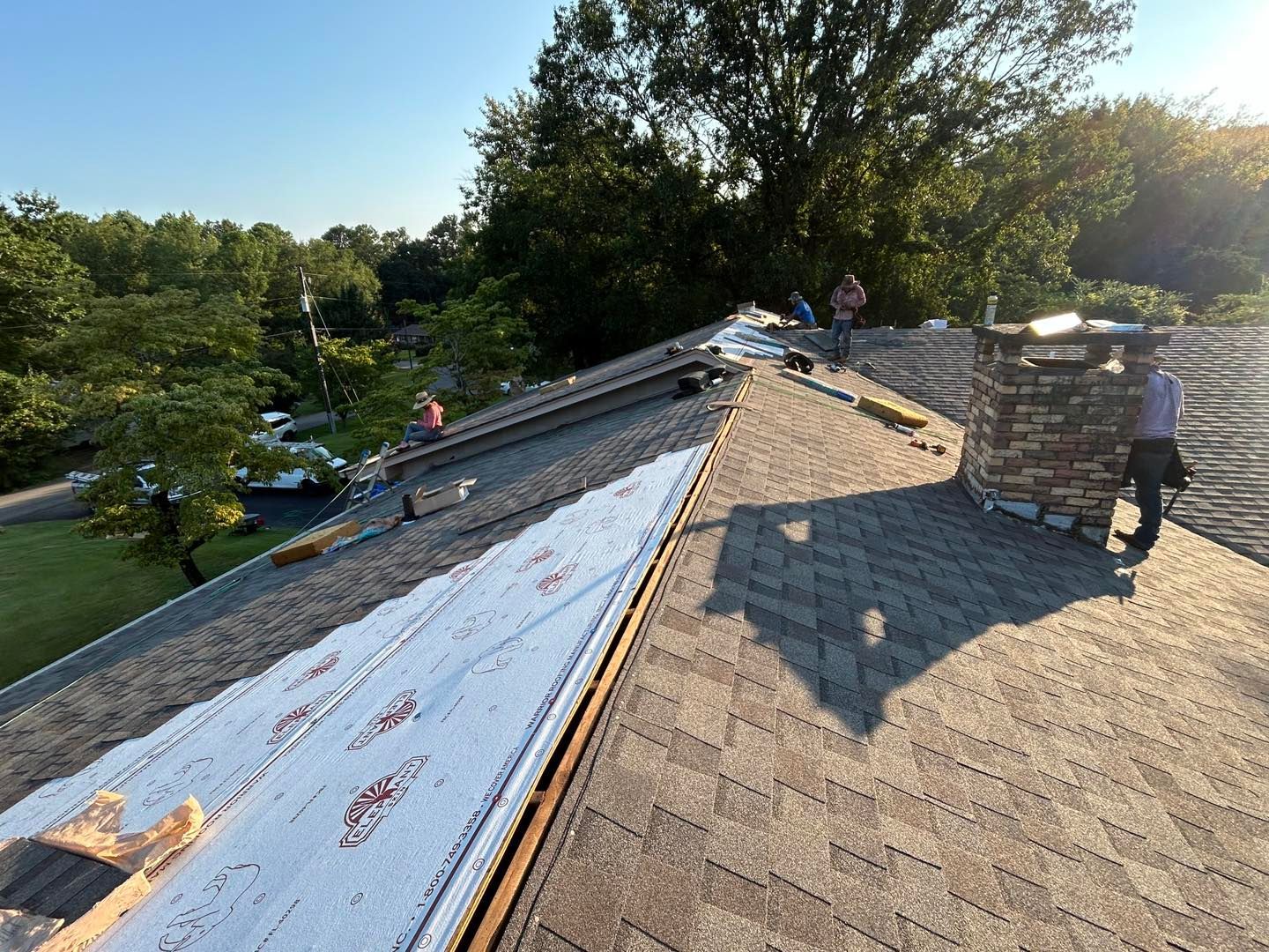 Roofers working on a residential roof, near a stone chimney, with trees in the background.