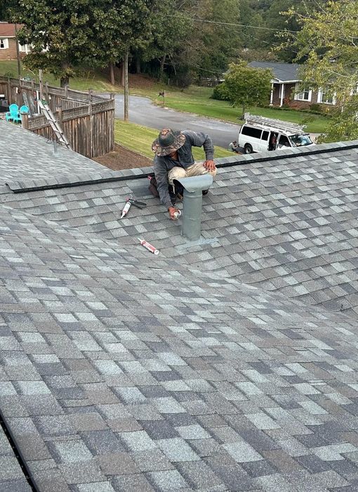 Roofer on a gray shingle roof, working around a metal vent pipe, with a yard and houses in the background.