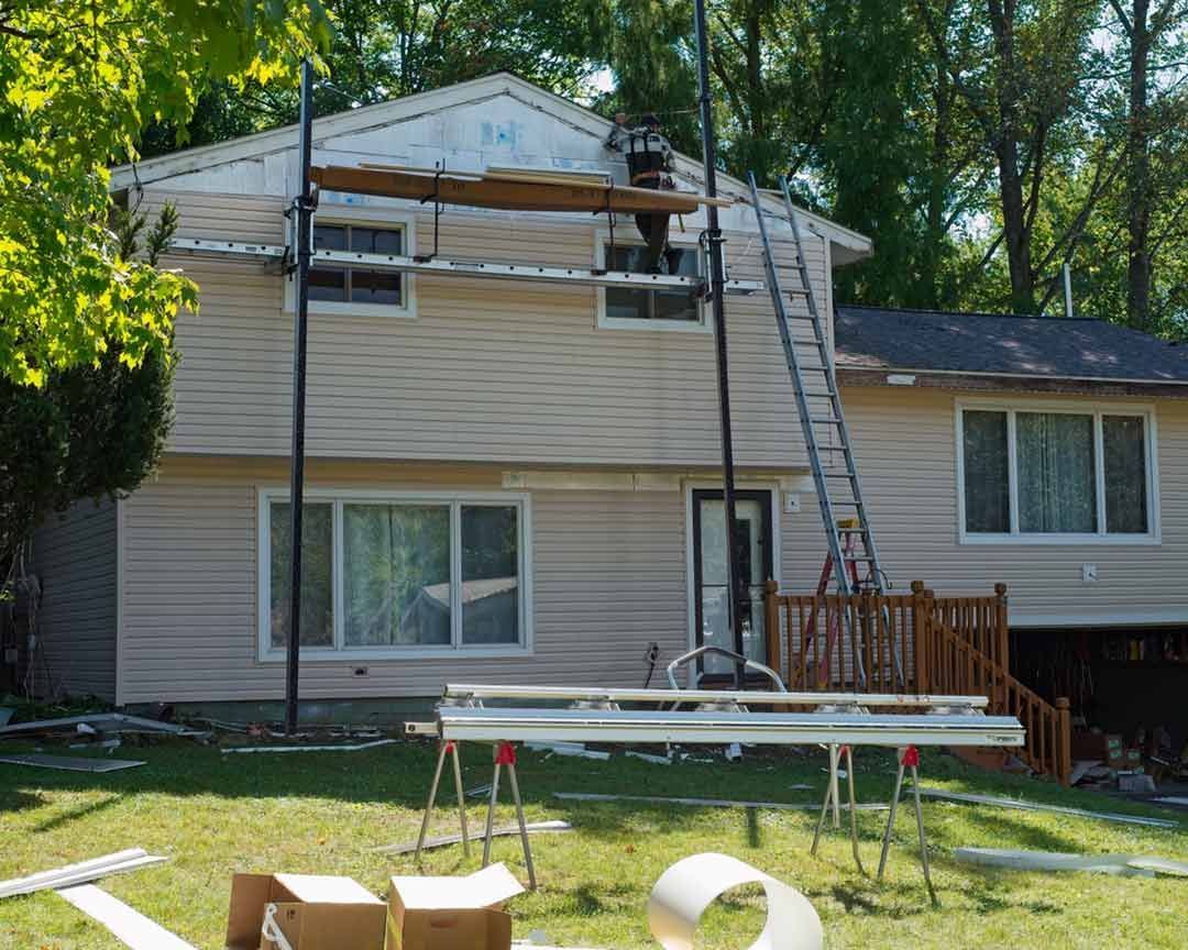 House undergoing siding replacement, scaffolding and ladder in place. Light tan siding, green lawn, sunny day.