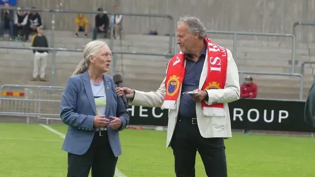 Två personer står på en fotbollsplan; en person i blå kavaj talar i en mikrofon som hålls av en person i vit jacka.