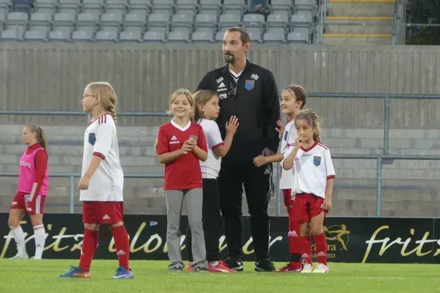 En tränare står på en fotbollsplan med flera barn i tröjor, med en spelare synlig till vänster.
