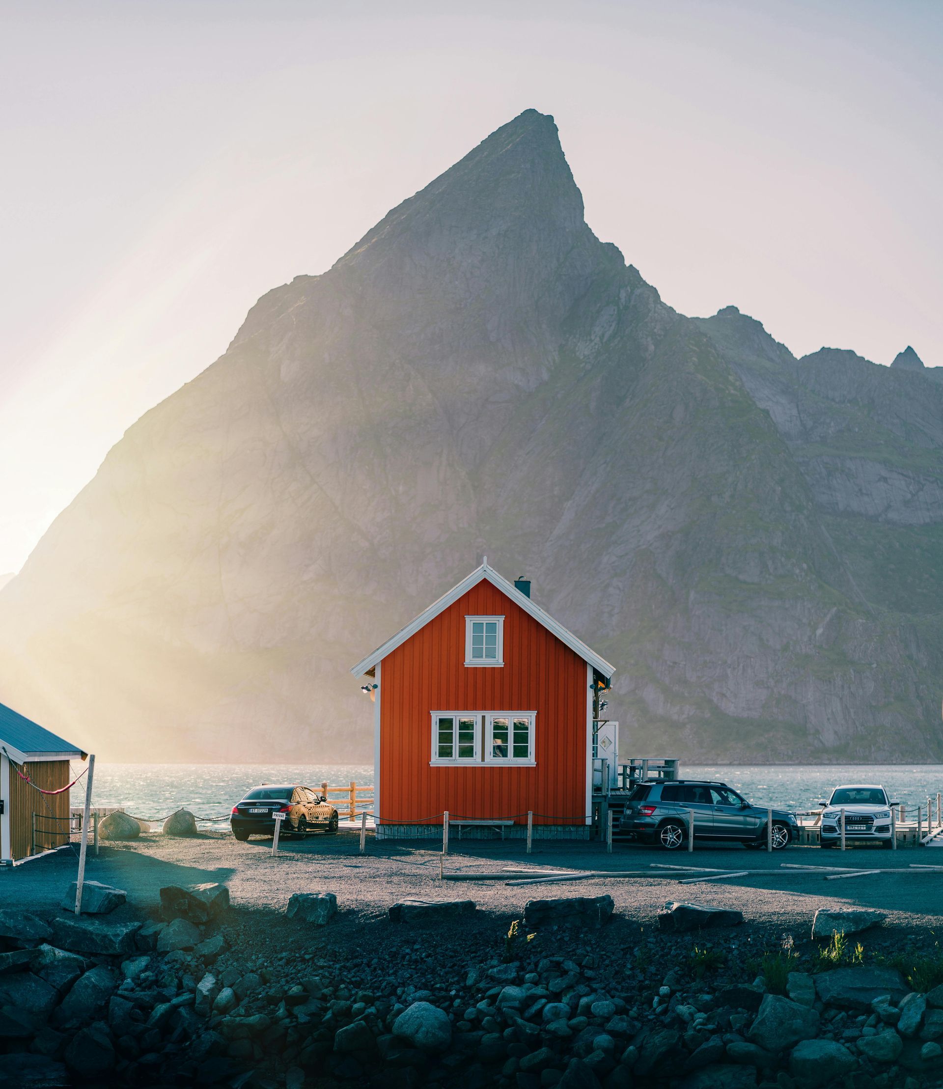 Rød hytte ved vann med fjell i bakgrunnen og parkerte biler.