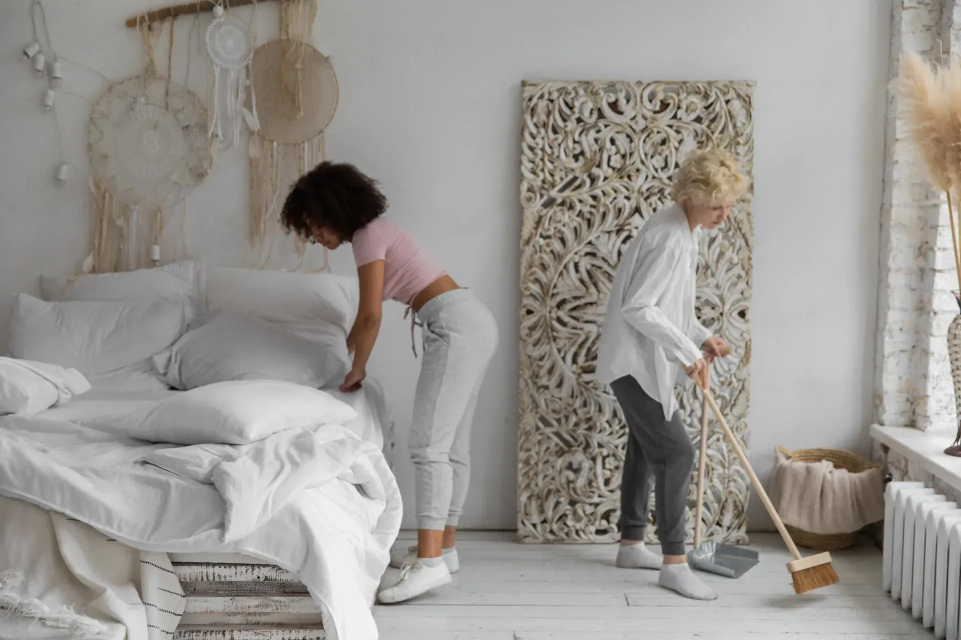 Two people tidying a bright bedroom, one making the bed and one sweeping the floor