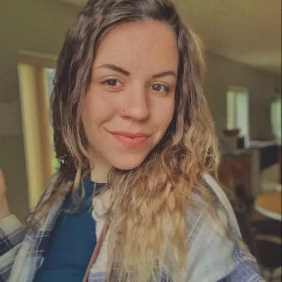 Smiling person indoors wearing a blue top and plaid shirt, with long wavy hair.
