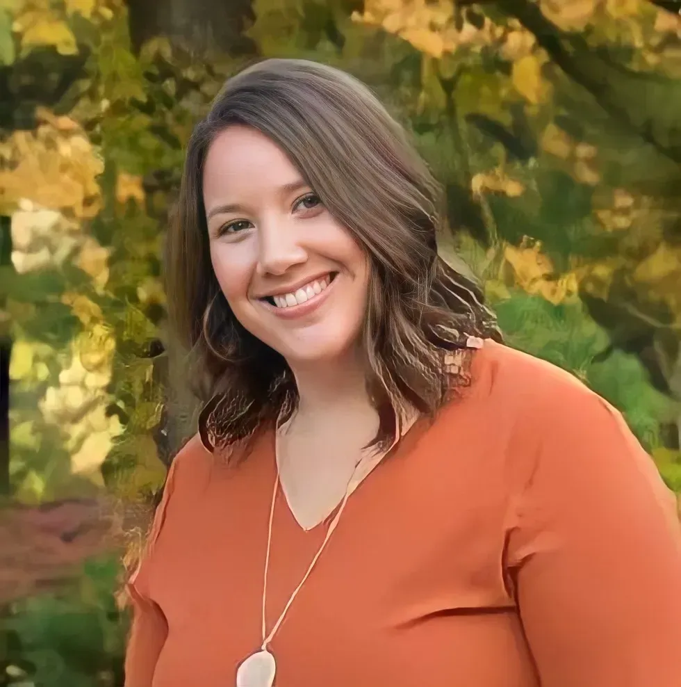 Smiling woman in an orange top standing outdoors with blurred autumn foliage in the background