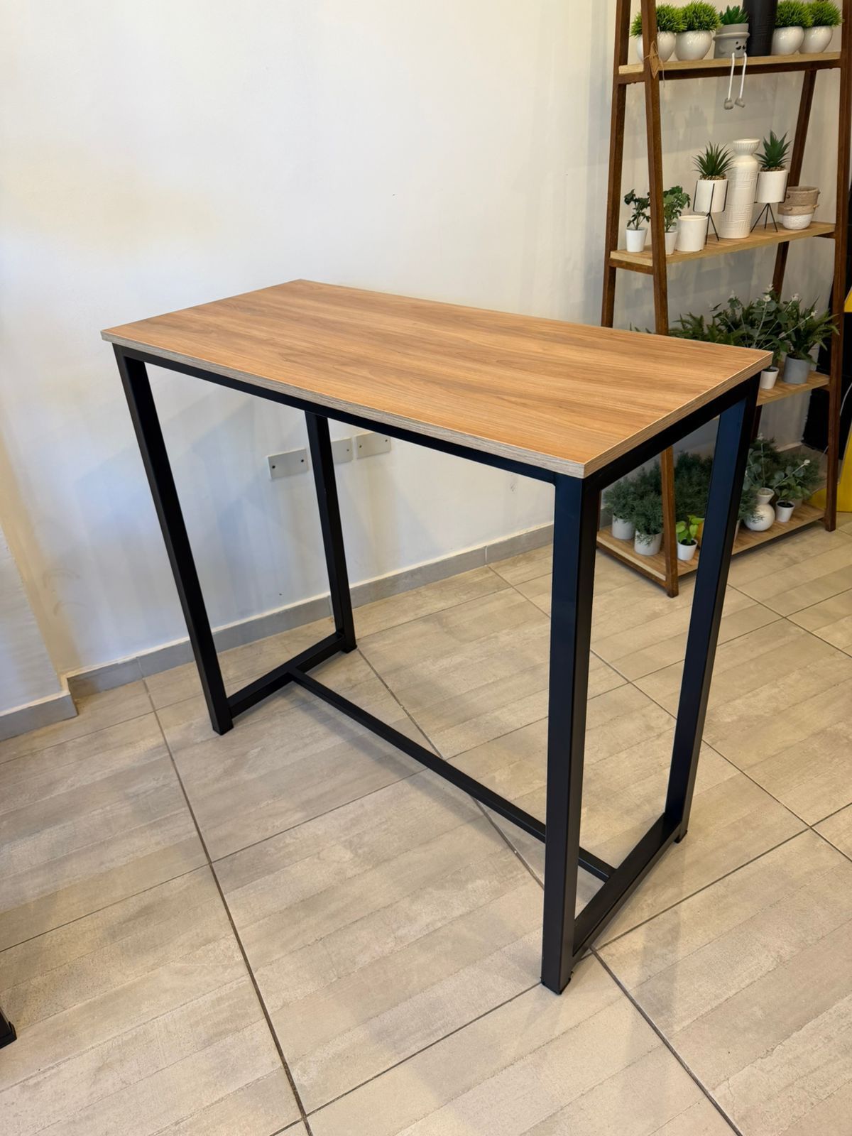 Mesa de metal negro con tablero de madera en una habitación alicatada, con estantes y plantas al fondo.