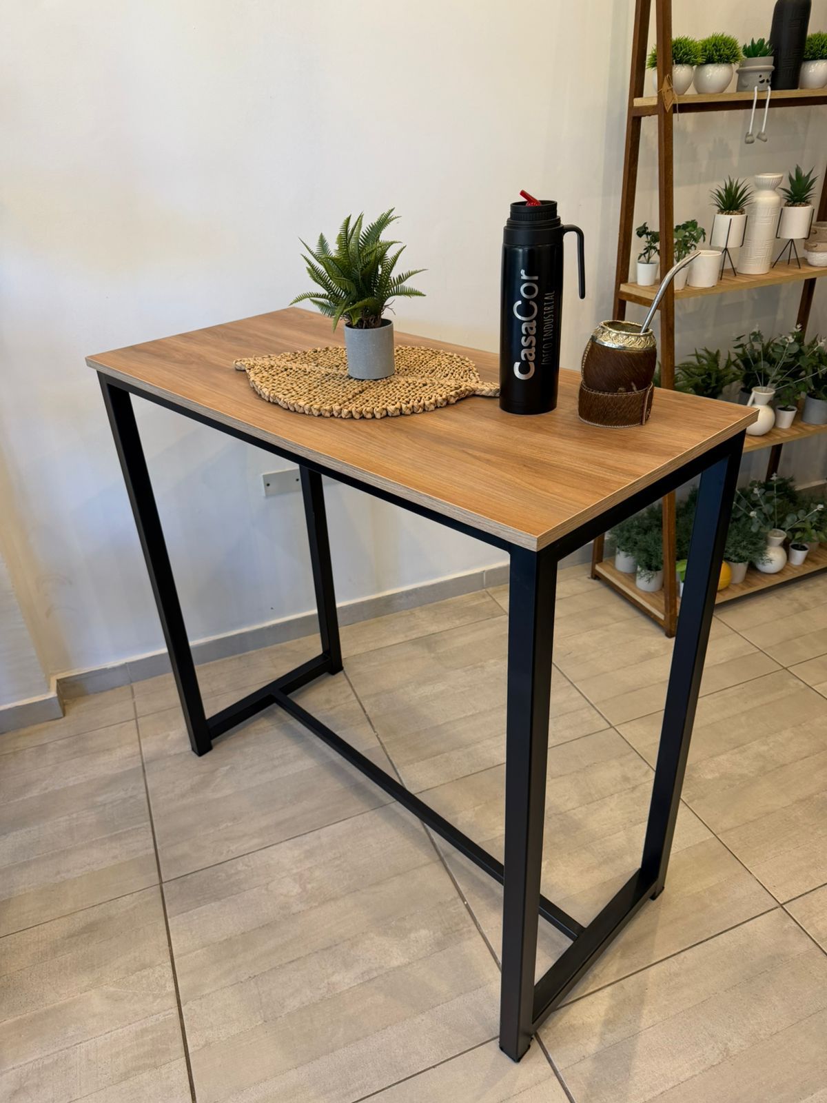 Mesa de metal negro con tablero de madera en una habitación alicatada, con estantes y plantas al fondo.