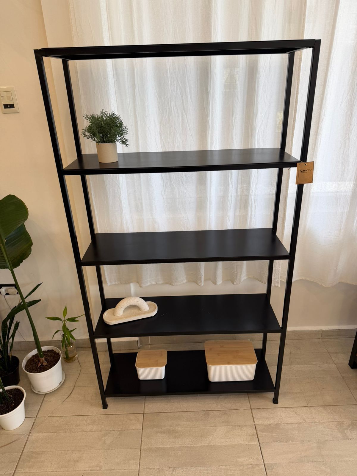 Estantería de metal negro con plantas en macetas, jabón y toallas en una habitación luminosa.