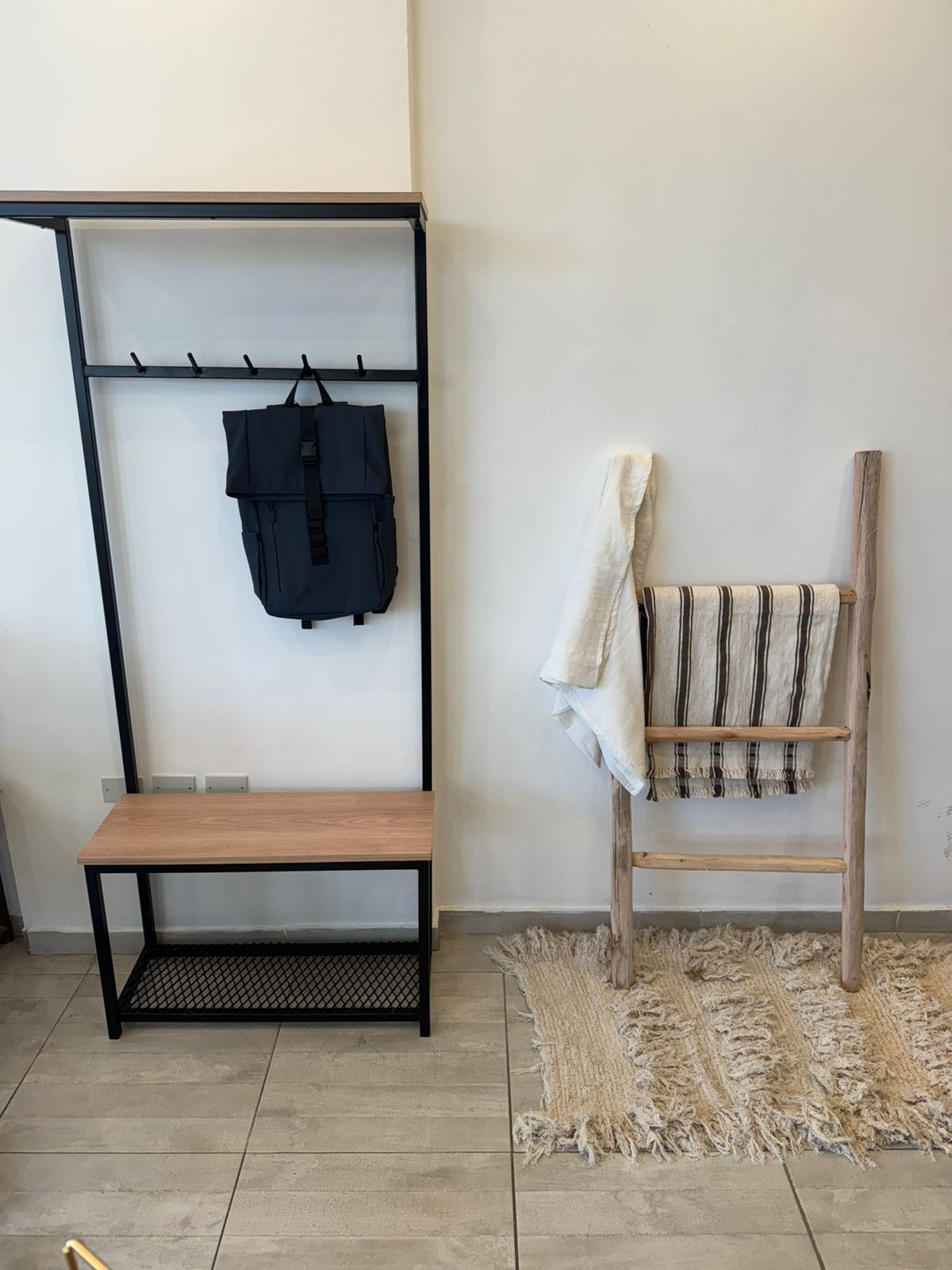 Entrada minimalista con perchero negro, banco y silla de madera sobre una alfombra tejida contra una pared blanca.