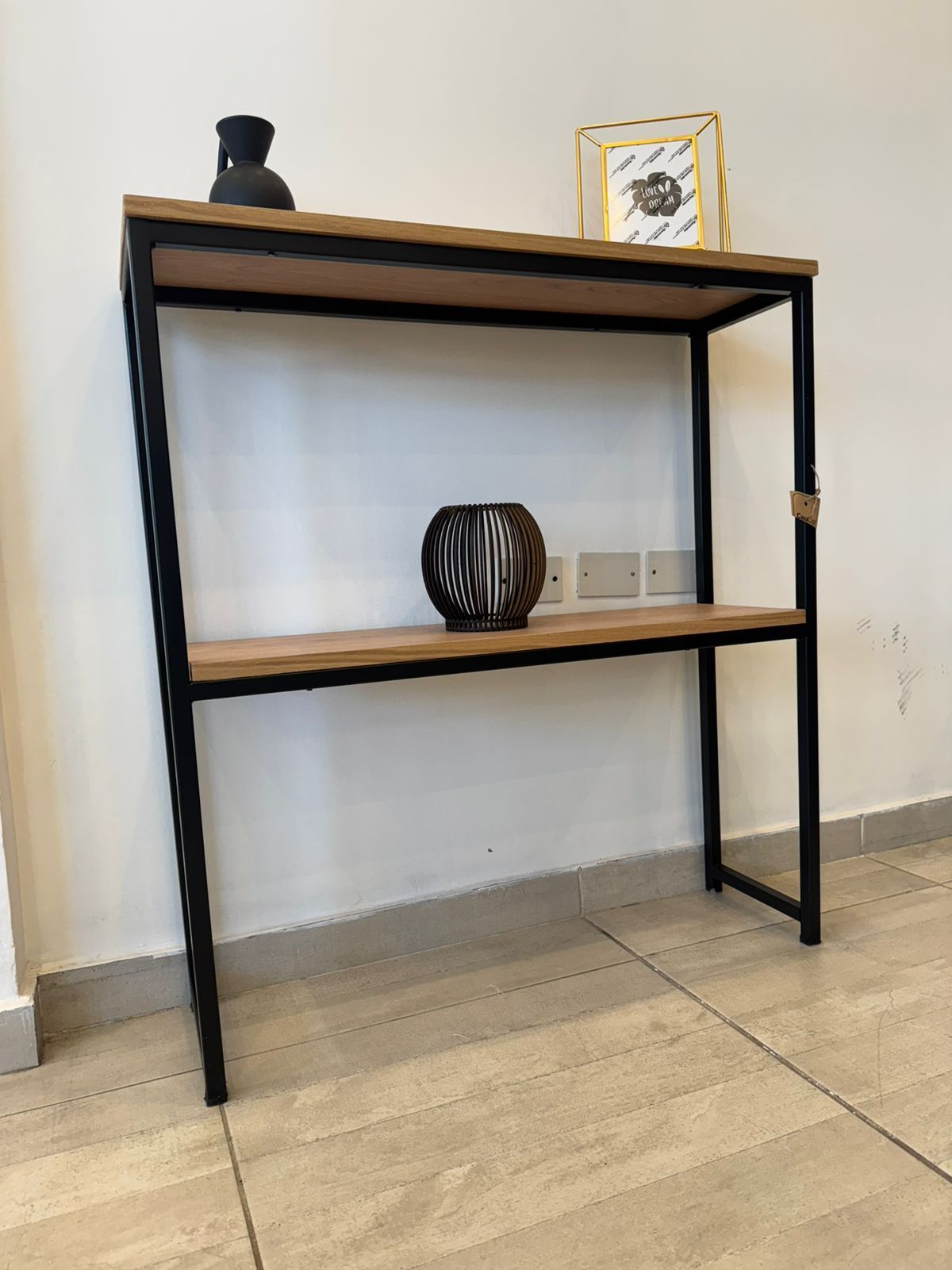 Mesa consola de madera con jarrón negro, foto enmarcada y esfera decorativa sobre una pared blanca.