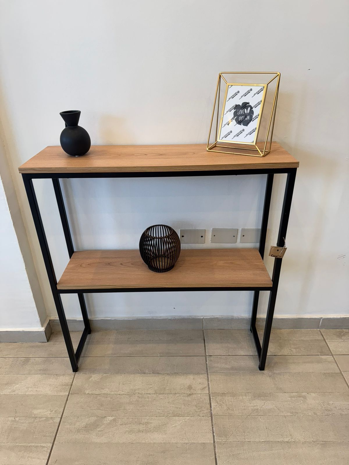 Mesa consola de madera con jarrón negro, foto enmarcada y esfera decorativa sobre una pared blanca.