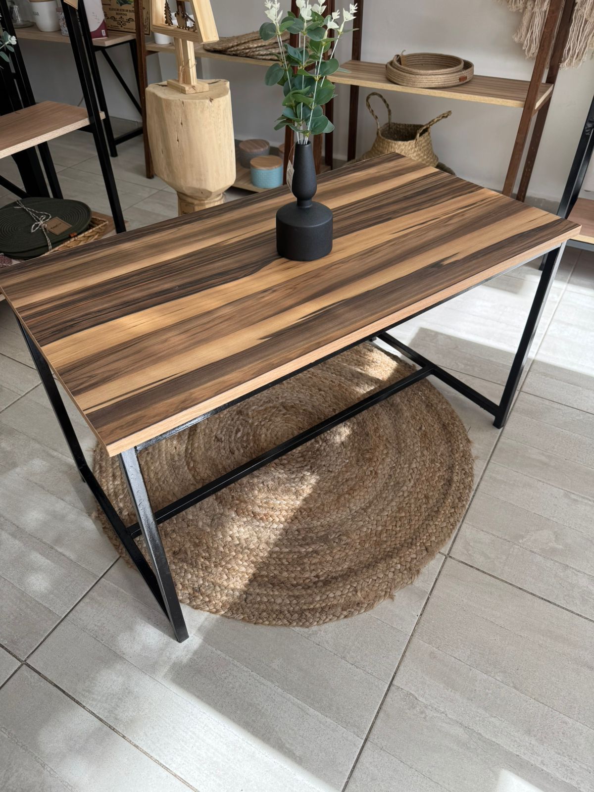 Mesa de madera con patas de metal negro sobre una alfombra tejida en una habitación luminosa.