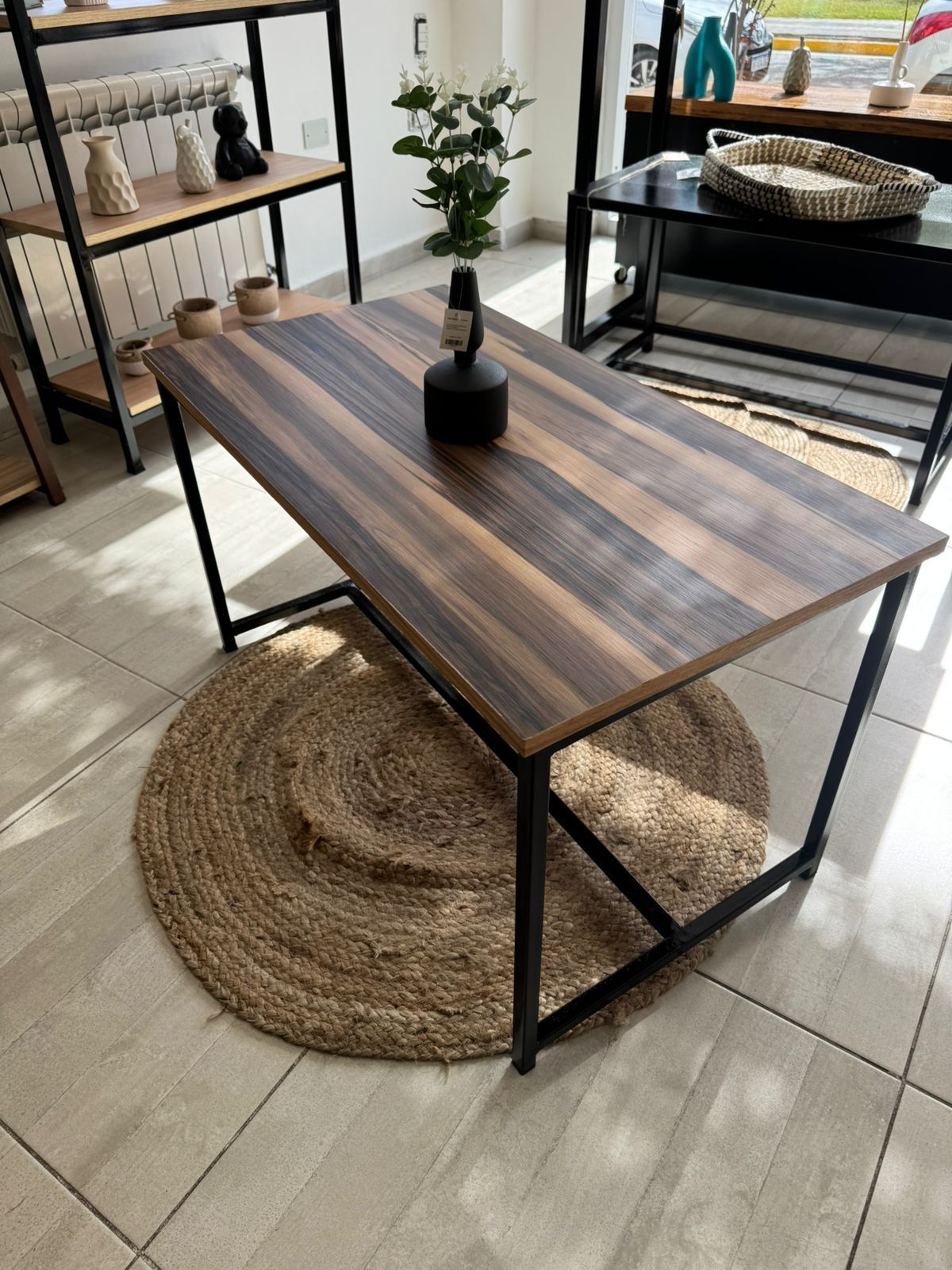 Mesa de madera con patas de metal negro sobre una alfombra tejida en una habitación luminosa.